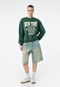 Zielony oversize'owy sweatshirt z nadrukiem "NEW YORK" w białym kolorze, w połączeniu z jasnoniebieskimi dżinsowymi szortami. Model nosi białe sneakersy i skarpetki.