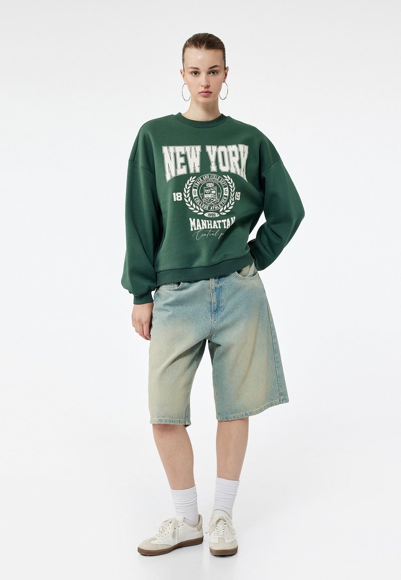 Zielony oversize'owy sweatshirt z nadrukiem "NEW YORK" w białym kolorze, w połączeniu z jasnoniebieskimi dżinsowymi szortami. Model nosi białe sneakersy i skarpetki.