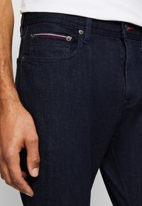 Gros plan sur la main d'une personne reposant à côté d'un jean denim bleu foncé avec une poche avant visible et des détails de coutures rouges, blanches et bleues.