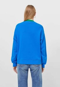 Sudadera azul con cuello redondo y ribete verde, de mangas largas y corte relajado, combinada con jeans de mezclilla azul claro.