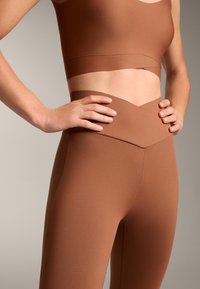 Barna aktív viselet szett, amely egy rövidített topból és magas derekú leggingsből áll, csehbán tervezéssel. Simább textúra és látható varrási részletek jellemzik.
