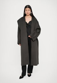 Dlouhý kabát z tmavě šedé vlněné směsi s oversized límcem a spadlými rameny, nošený přes černý top s cowl krkem a černé kalhoty.