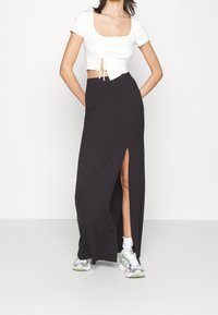 Femme portant un crop top blanc à manches courtes, une jupe longue noire fendue, des chaussettes blanches et des baskets argentées, debout devant un fond blanc uni.