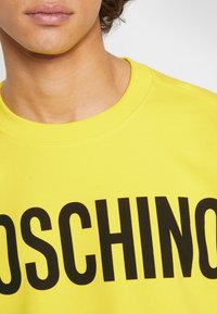 Zbliżenie osoby noszącej jasnosłoneczną żółtą bluzę z dużym czarnym napisem "MOSCHINO" na piersi.