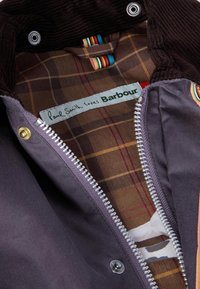 Casaco castanho-escuro e roxo com colarinho de cortiça, apresentando um forro interior estampado e um fecho de correr prateado. Etiqueta de marca visível.