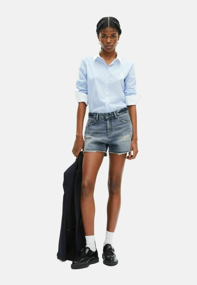 Jeune femme portant une chemise bleu clair, un short en jean, des chaussettes blanches et des chaussures noires, tenant une veste sombre contre un fond uni.