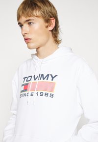 Fehér kapucnis pulóver állítható zsinórral, grafikai nyomattal, melyen a "TOMMY" felirat és piros, kék, valamint sárga csíkos minta található.