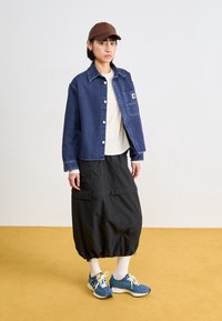 Veste en denim avec fermeture à boutons, poche poitrine ; superposée sur une chemise crème. Jupe noire avec ourlet froncé ; chaussures de sport bleues avec lacets blancs.