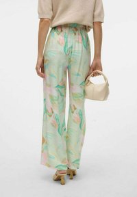 Pantaloni leggeri e fluide in un verde pastello con motivi floreali. A vita alta e con gambe ampie, abbinati a un top e una borsa beige.