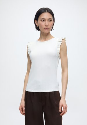 Kvinde iført en ærmeløs hvid top med flæser på skuldrene og mørkebrune viddebukser, stående foran en ensfarvet hvid baggrund.