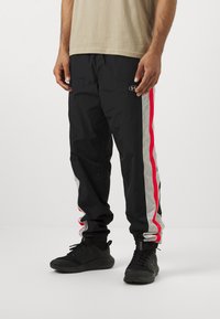 Black athletic pants with red and grey side stripes, made of lightweight material. Pairing with black sneakers and a beige T-shirt.