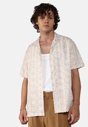 Camisa de manga corta en beige claro con un patrón geométrico, que cuenta con un cuello de campamento y un frente abierto. Lleva puesta una camiseta blanca debajo.