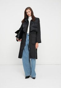 Zwarte puffer vest over een lange zwarte jas met een trekkoord in de taille, gecombineerd met lichtblauwe wijde jeans en een zwarte clutch handtas.