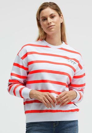 Woman wearing white long-sleeve shirt with red horizontal stripes and "Voyage" text on chest, paired with blue jeans, standing against gray background.