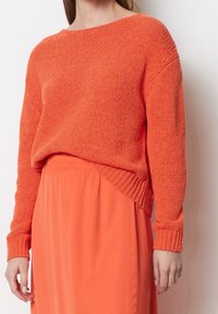 Pomarańczowy sweter z dzianiny o luźnym kroju i szerokim dekolcie. Rękawy i dół wykończone ściągaczami, w połączeniu z pasującą pomarańczową spódnicą. Gładka struktura.