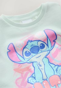 Mintgrøn sweatshirt med rund hals, prydet med en farverig grafik af en tegneseriefigur med store ører og udtryksfulde øjne.