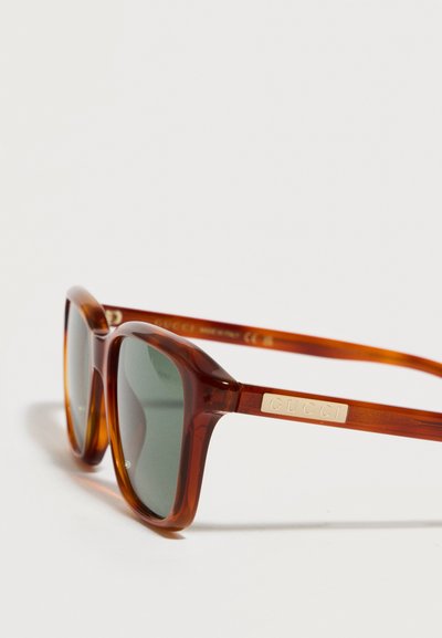 Lunettes de soleil Gucci translucides marron avec des verres rectangulaires teintés verts et un logo de la marque en or sur la branche.