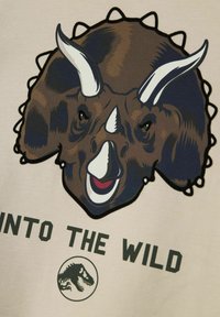 Brun t-shirt med en grafik af et dinosaurhoved med hvide horn. Teksten "INTO THE WILD" står under designet. Cremefarvet baggrund.