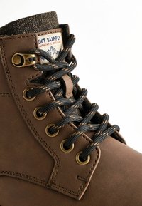 Brown leather boot with black and orange laces, metal eyelets, and a fabric collar. Label features blue text and mountain design.