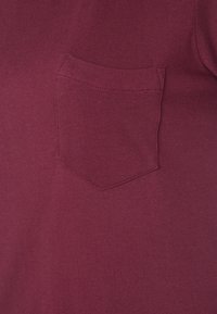 Burgundská košile s krátkým rukávem a kapsou na prsou. Tkanina je jemná a texturovaná, s decentním prošitím kolem kapsy.