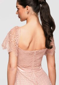 Femme aux cheveux foncés attachés en queue de cheval, portant une robe en dentelle rose avec des manches en dentelle transparentes, une ceinture en satin et une fermeture éclair au dos.