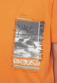 Oranje katoenen T-shirt met een grijze grafiek van de zon, golven en tekstaccenten. Het ontwerp benadrukt een strandthema met een textuureffect.