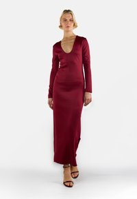 Kobieta w długiej, bordowej wieczorowej sukni z głębokim dekoltem w kształcie litery V, w czarnych sandałach na kostkę, stojąca na białym tle.