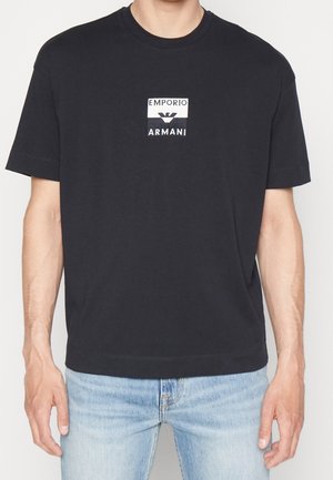 Czarna bawełniana koszulka z kwadratowym, białym logo z napisem 'Emporio' i 'Armani'. Krótkie rękawy, okrągły dekolt i luźny krój.