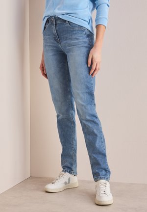 Jean en denim bleu clair avec une coupe slim, présentant un design classique à cinq poches et un léger délavage. Accompagné de baskets blanches.