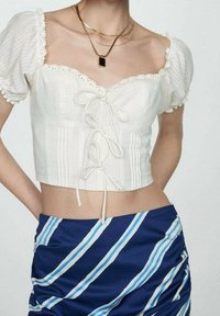 Haut blanc court avec un design smocké et une bordure en dentelle ; comprend des manches courtes bouffantes et un lien à l'avant. Associé à une jupe bleue à rayures.