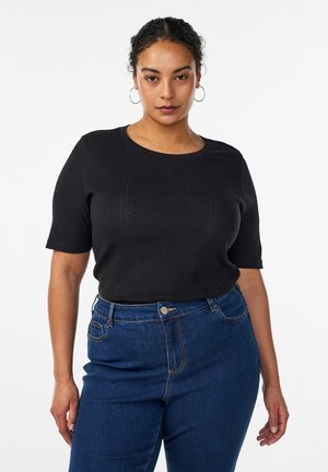 Vrouw met krullend haar die hoepeloorbellen draagt, een zwart top met korte mouwen en blauwe jeans met hoge taille, staand tegen een witte achtergrond.