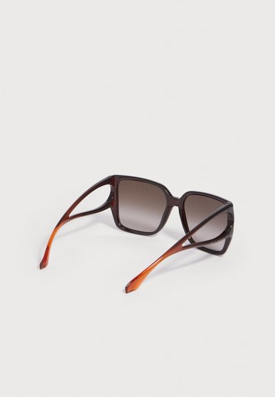 Lunettes de soleil carrées avec verres dégradés foncés et montures translucides marron, vues de dos sur une surface blanche.