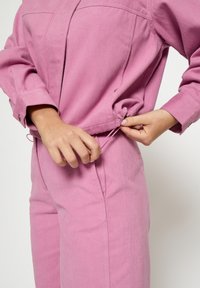 Rozā, lina maisījuma tops ar pogu aizdari un aukliņu apakšmalā. Valkātājs pielāgo aukliņu pie vidukļa, uzrādot vaļīgu piegulumu.