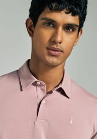 Vaaleanpunainen polo-paitaja, joka on valmistettu pehmeästä kankaasta. Siinä on kauluspaita, kolmen napin kaulus ja brodeerattu peura-logo vasemmassa rinnassa.