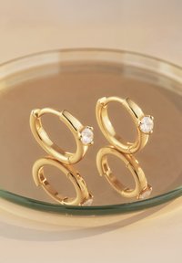 Guldörhängen med cirkelform och polerad finish, dekorerade med en enda klar ädelsten. Rund form med slät yta, placerade på en reflekterande yta.