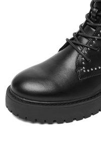 Bota de tobillo de cuero negro con punta redondeada, superficie texturizada, parte delantera con cordones y tachuelas decorativas plateadas a lo largo del costado. Suela negra gruesa.