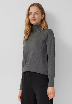 Woman with long blonde hair wearing a grey turtleneck sweater and black pants, standing against a plain light background.