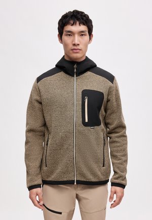 Ung mand iført en beige og sort tekstureret lynlåstrøje med en sort brystlomme og beige bukser mod en ensfarvet hvid baggrund.