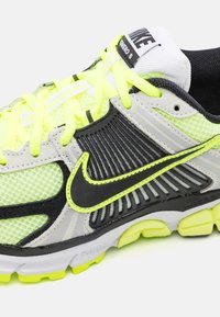 Nike löparsko med ett mesh-överkär som har svarta, grå och neon gula detaljer, texturerade sidor och dämpad sula. Ljusa snören och logotyp synliga.