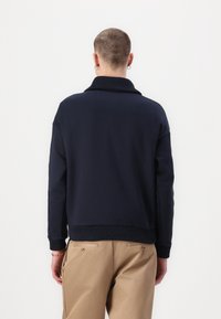 Sudadera azul marino con un cuello alto, puños y dobladillo acanalados. Se lleva con pantalones caqui, mostrando un diseño limpio y sencillo.