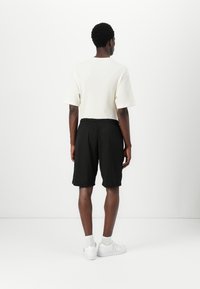 Short-sleeve, ribbed white shirt paired with loose-fitting black mesh shorts; model wears white sneakers and sits against a plain backdrop.