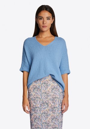 Blauer, gestrickter V-Ausschnitt-Pullover mit kurzen Ärmeln, der eine strukturierte Oberfläche aufweist. Kombiniert mit einem Rock im floralen Muster in sanften Pastellfarben.