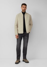 Gestepptes beige Bomberjacke mit Reißverschluss vorne, kombiniert mit dunklen Jeans und braunen Schuhen. Die Jacke hat einen gerippten Kragen und eine strukturierte Oberfläche.