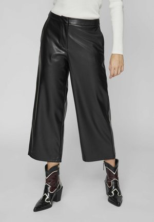 Frau trägt schwarze Lederhose mit weitem Bein, weißes geripptes Langarmshirt und schwarz-bordeauxfarbene spitze Cowboy-Stiefel mit weißen Details.