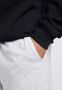 Mano di una persona nella tasca di pantaloni della tuta grigi che indossa una maglia nera a maniche lunghe con polsini risvoltati su uno sfondo semplice.