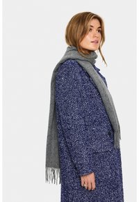 Kvinde med lyst brunt hår iført en blå hørskindjakke og grå frynset tørklæde, stående sidelæns foran en ensfarvet hvid baggrund.