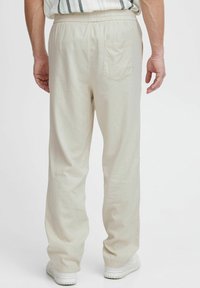 Man in losse beige broek met elastische tailleband en achterzak, gecombineerd met witte sneakers, staand tegen een witte achtergrond.