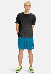 Sort kortærmet atletisk t-shirt, paired med teal shorts med Fila-logoet. Modellen har grønne og sorte sneakers på.