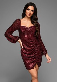 Robe rouge pailletée avec de longues manches bouffantes, décolleté en cœur et fronces sur les côtés. Le tissu a une apparence glossy et texturée.