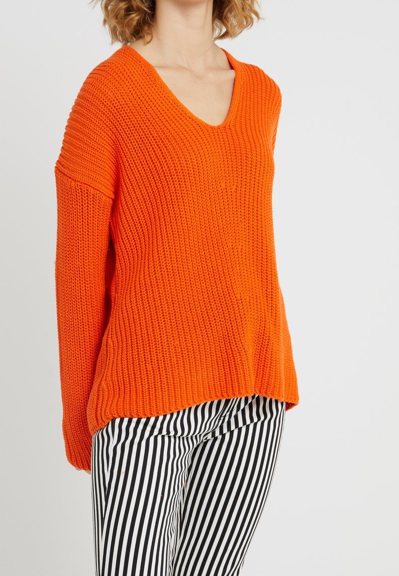 Femme portant un pull en grosse maille orange vif à col en V et un pantalon à rayures verticales noires et blanches sur un fond uni.